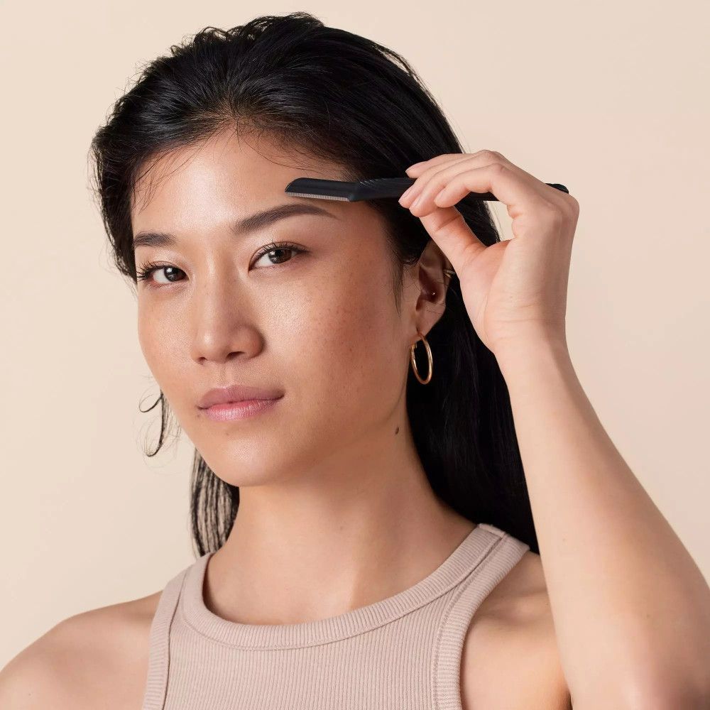 Une personne tient un rasoir à sourcils. Manche noir, lame argentée. Personne à la peau claire et haut beige.