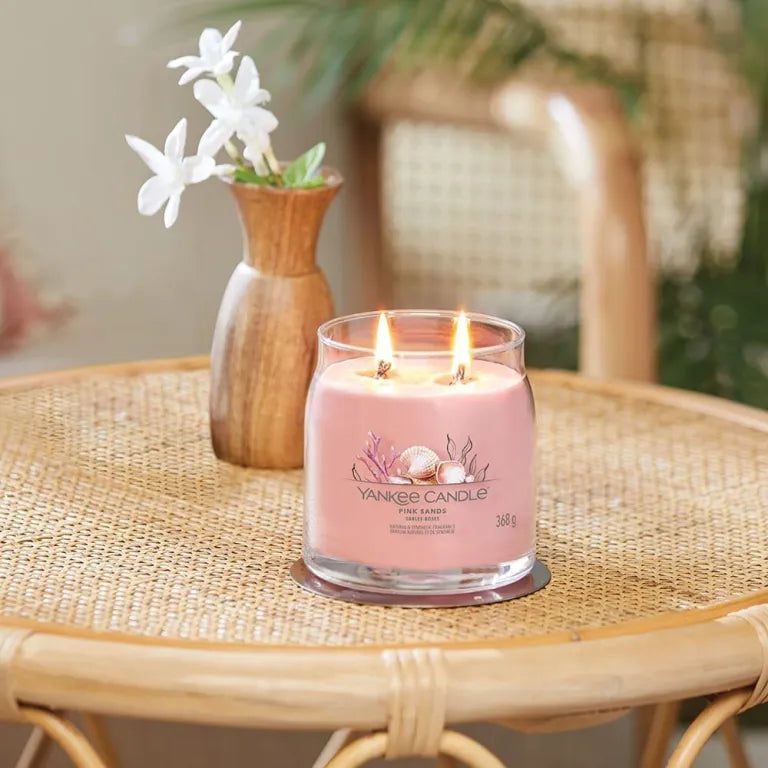 Bougie rose Yankee Candle Pink Sands sur une table. Deux mèches allumées. Vase avec fleurs en arrière-plan.