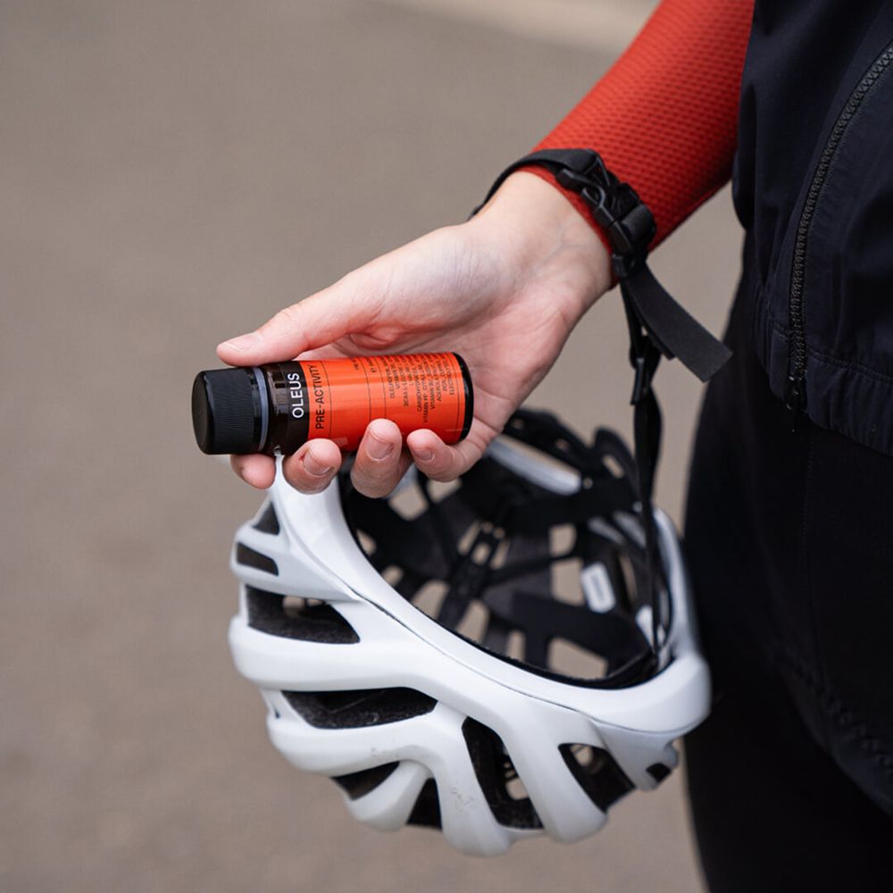 Hand houdt een fles OLEUS vast. Daarnaast een fietshelm. Rode mouw.