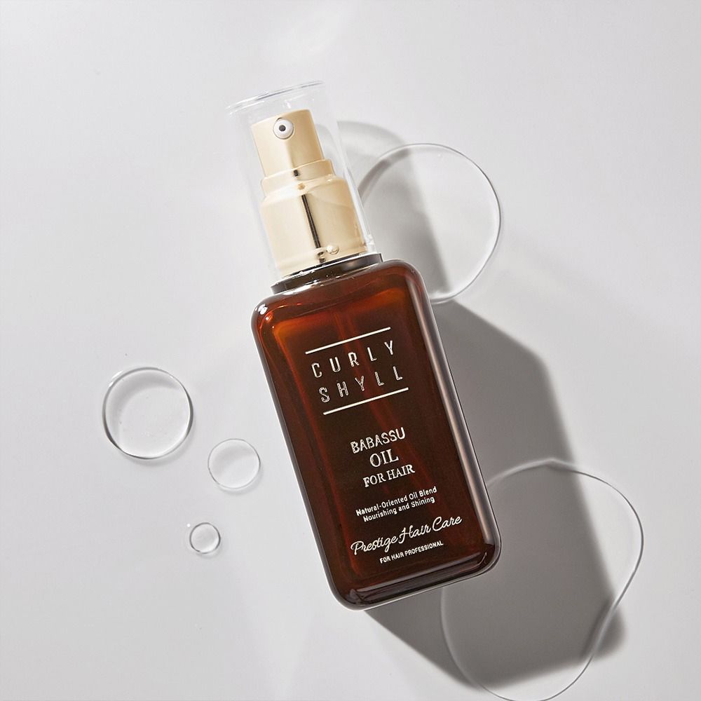 Bruine fles met gouden verstuiver. Opschrift: CURLY SHYLL, Babassu Oil for Hair. Omringd door waterdruppels.