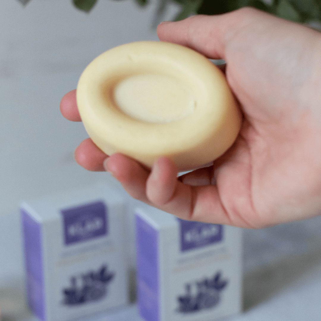 Hand houdt ovale, vaste body butter vast. Lichtbeige kleur, concave verdieping. Op de achtergrond twee verpakkingen met productnaam: Arganolie & Vijg.