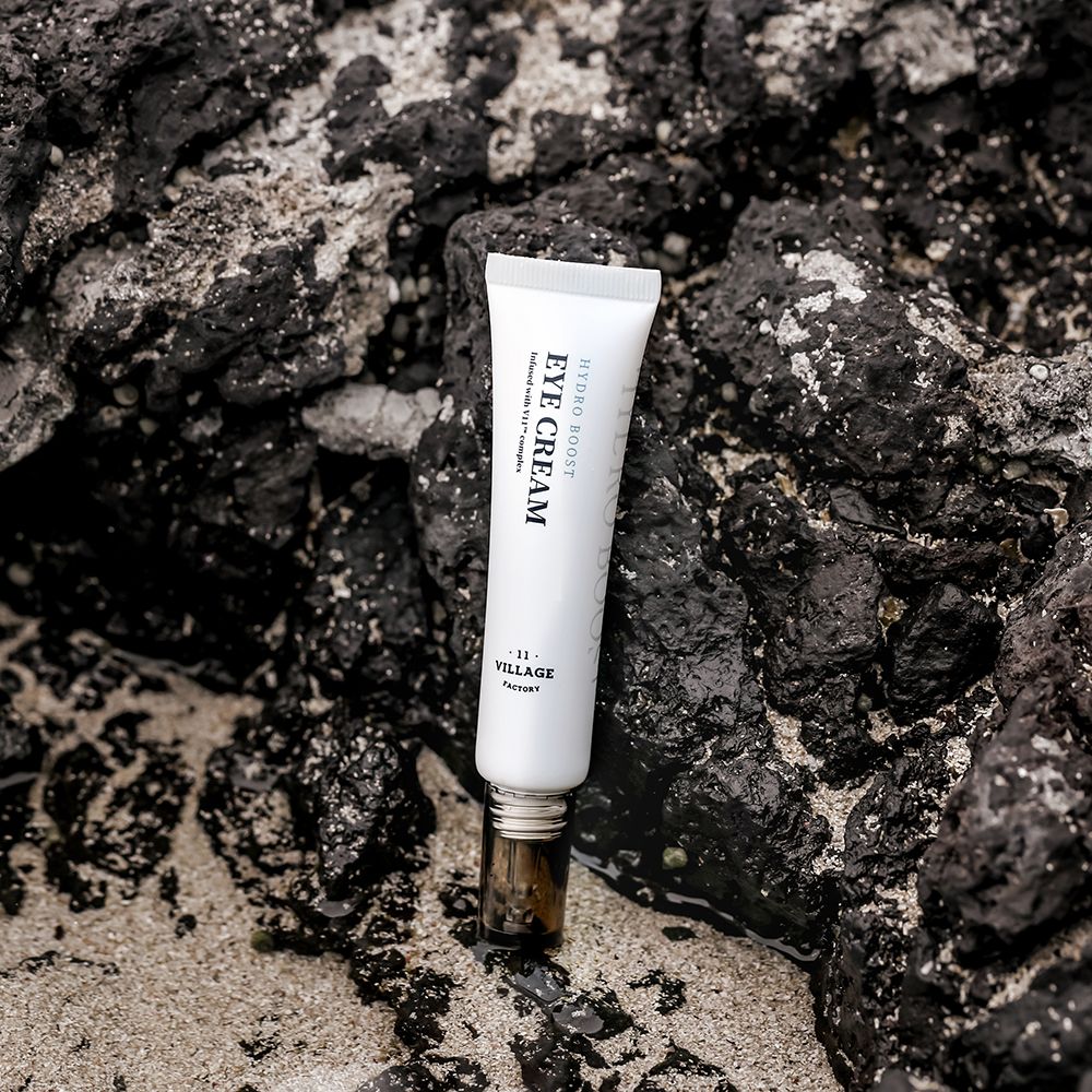 Witte tube op donkere stenen. Zwarte tekst: EYE CREAM, HYDRO BOOST, VILLAGE 11 FACTORY. Bruine sluiting.