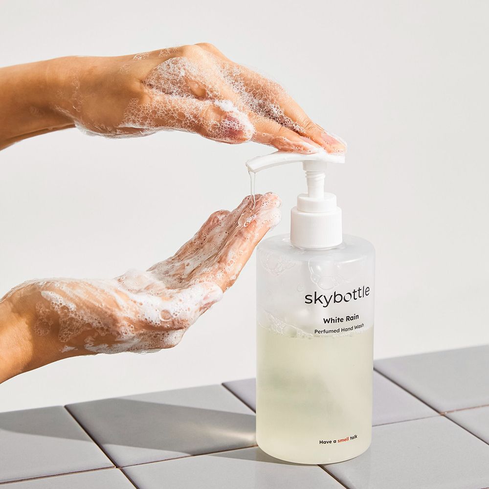 Handen die vloeibare zeep ontvangen uit een fles met witte pomp. Opschrift: skybottle, White Rain, Perfumed Hand Wash.