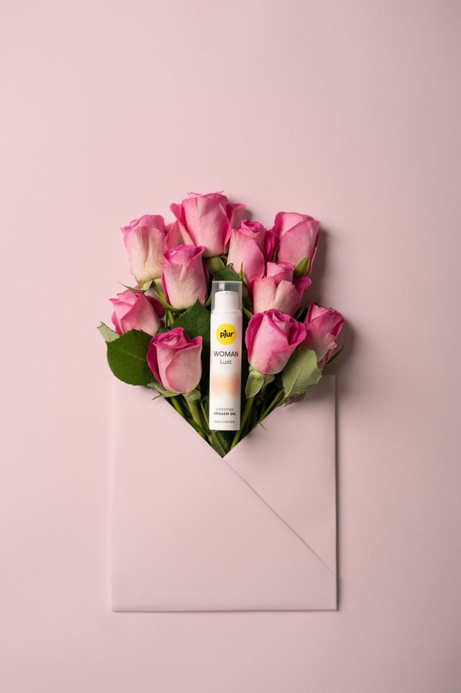 Flacon avec logo jaune, entouré de roses dans une enveloppe. Texte: WOMAN Lust, Vibrating Orgasm Gel. Fond rose.