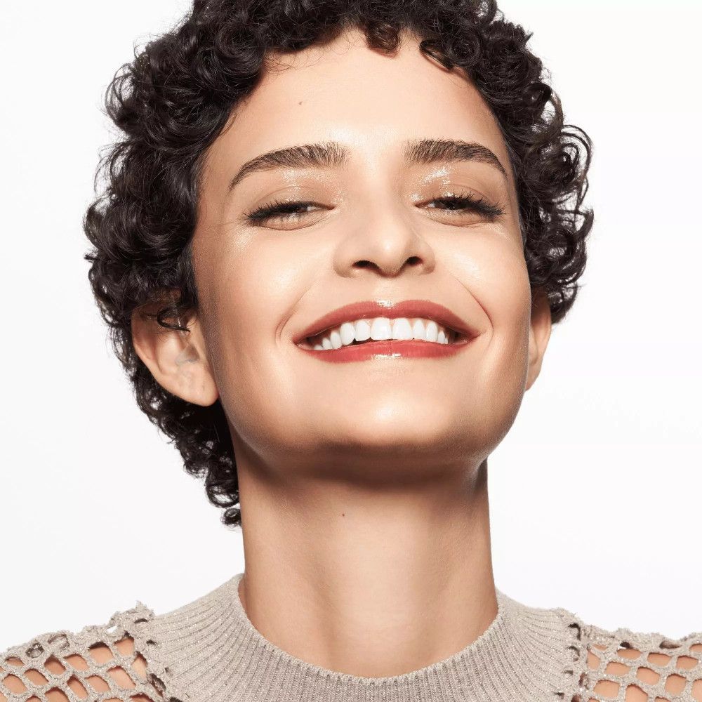 Femme aux boucles, portant du baume à lèvres. Sourit. Teint clair, lèvres brun clair.