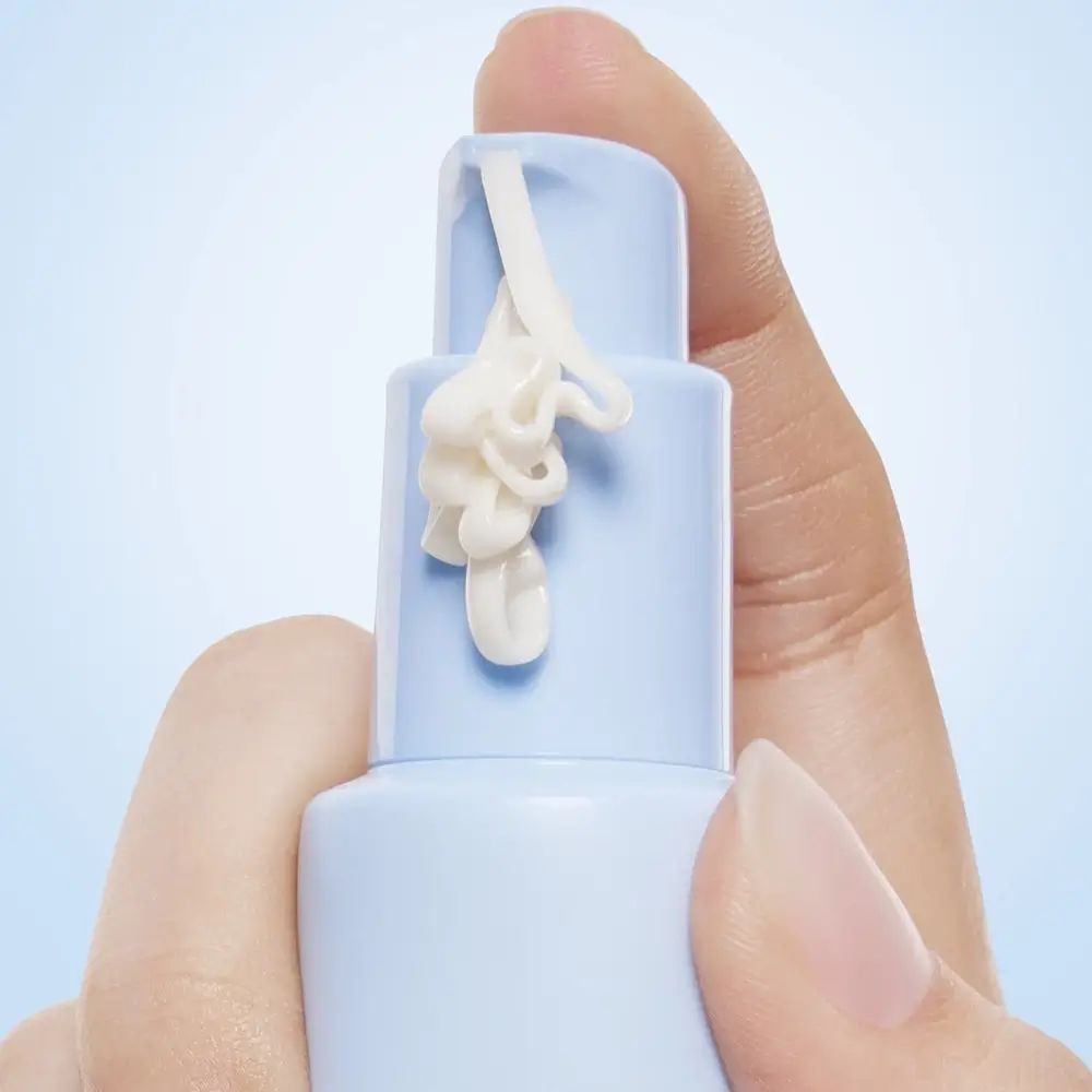 Close-up van een hand die een lichtblauwe tube vasthoudt. Crème wordt uit de opening geperst. Productnaam niet zichtbaar.