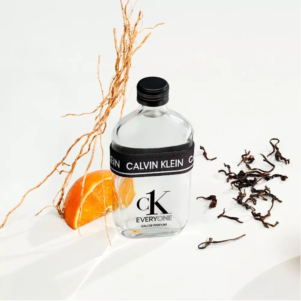Transparante glazen fles, zwarte dop en band met Calvin Klein. Tekst: ck EVERYONE. Met sinaasappel en takken.
