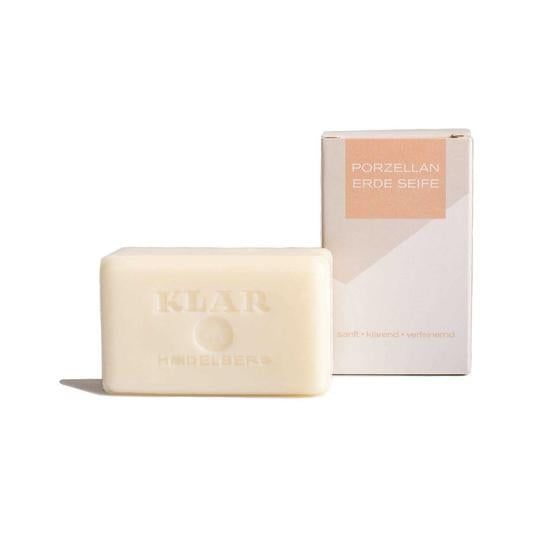 Savon à côté d'un emballage ouvert. Savon avec l'inscription KLAR. Emballage avec l'inscription PORZELLAN ERDE SEIFE. Texte en dessous : doux, clarifiant, affinant.