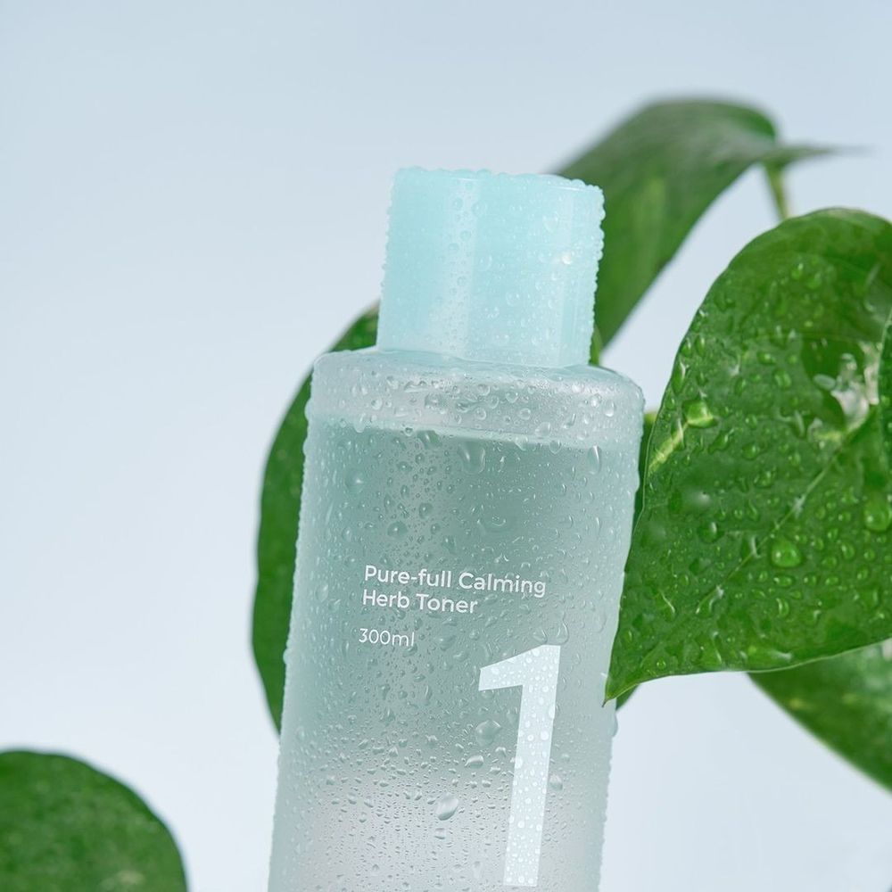 Flacon de toner transparent. Inscription: Pure-full Calming Herb Toner, 1, 300ml. Gouttes d'eau et feuilles vertes en arrière-plan.
