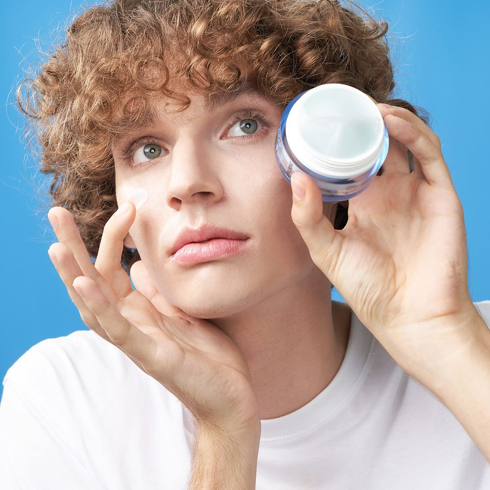 Un homme tient un pot de crème. Il porte un t-shirt blanc. Il touche son visage avec la crème.