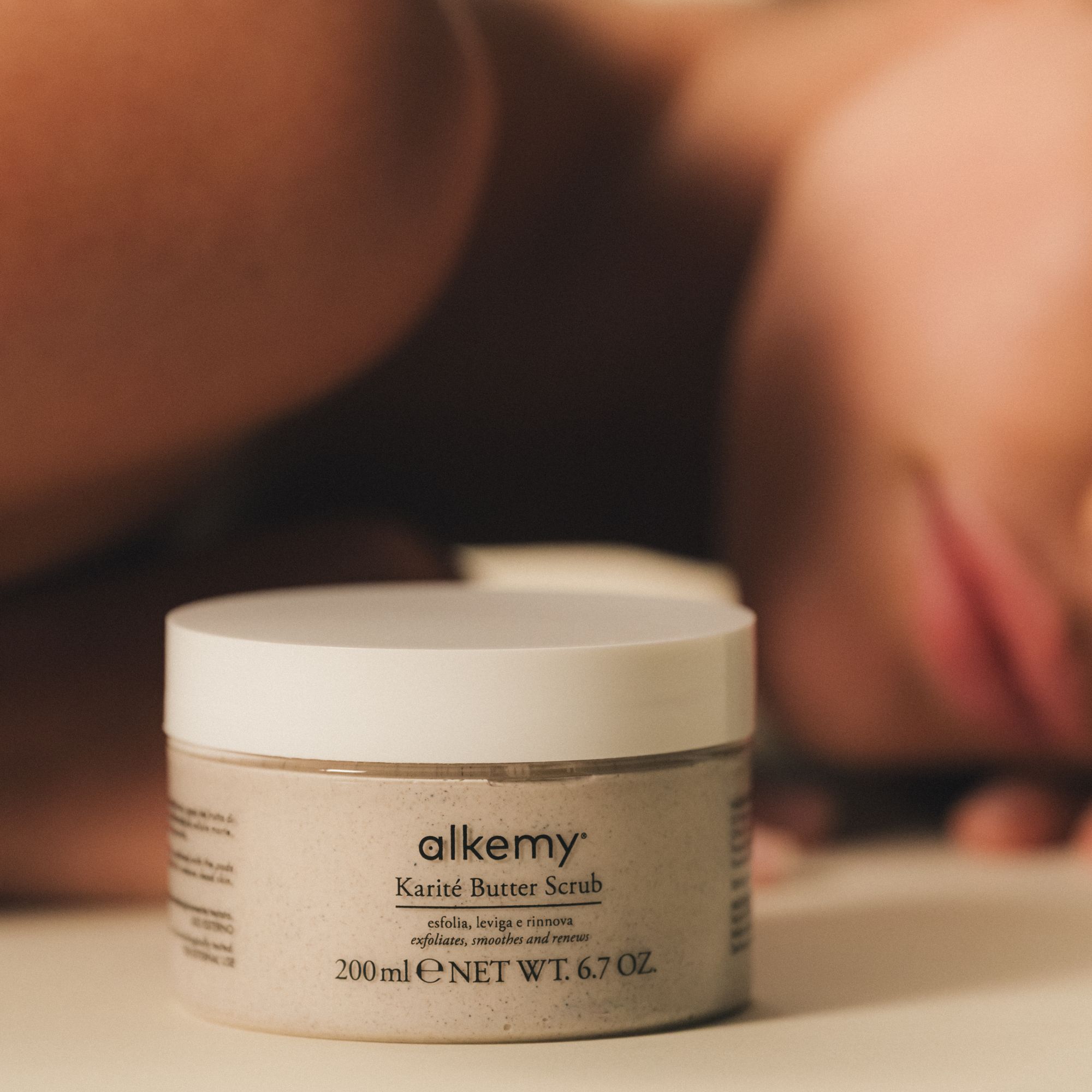 Gommage corps Alkemy Karité Butter Scrub dans un pot, à côté d'une personne allongée.