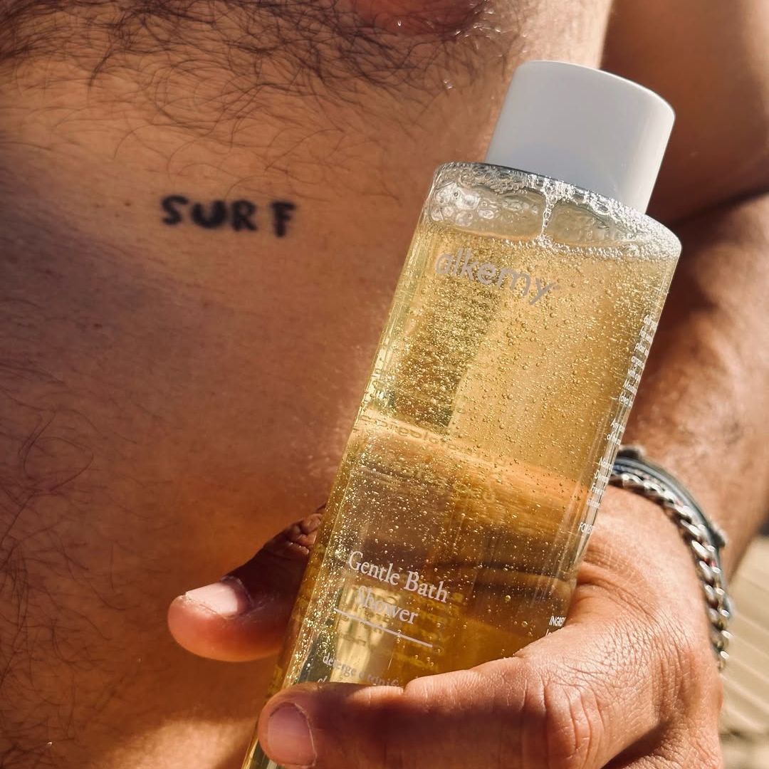 Gel douche Alkemy dans une bouteille transparente, tenu par une main devant une peau tatouée.