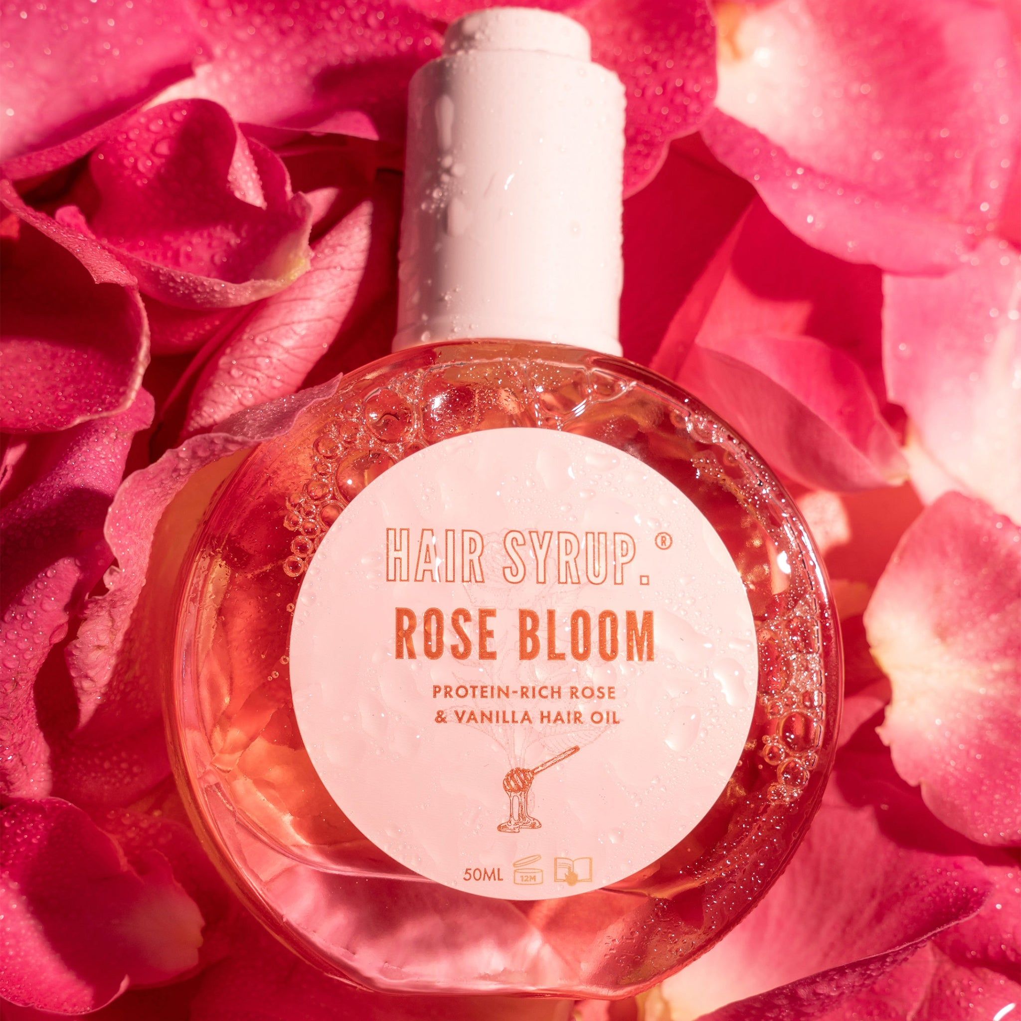 Ronde fles met witte dop, omringd door rozenblaadjes. Opschrift: Hair Syrup Rose Bloom. Eiwitrijke rozen- en vanille-haarolie.