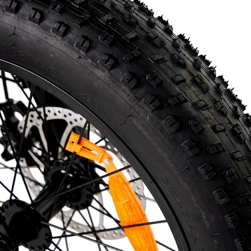 Gros plan sur un pneu de vélo noir avec une bande de roulement rugueuse. Frein à disque et roue à rayons visibles. Réflecteur orange.