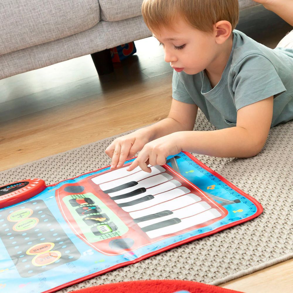 Un enfant est allongé sur un tapis et joue sur un tapis musical avec des touches de piano. Un bouton rouge est visible. Un canapé est en arrière-plan.
