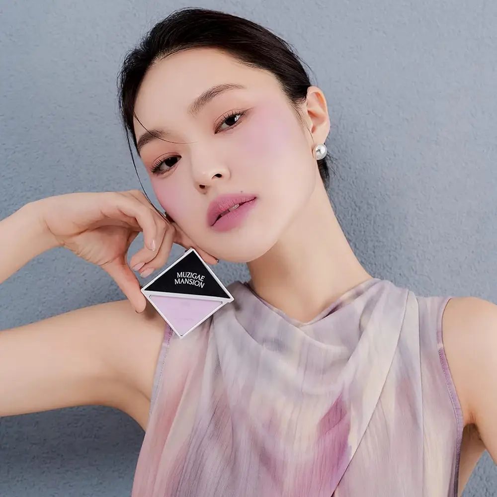 Vrouw houdt vierkante rougeverpakking vast. Zwart deksel met MUZIGAE MANSION. Roze rouge. Vrouw draagt rouge op wangen.