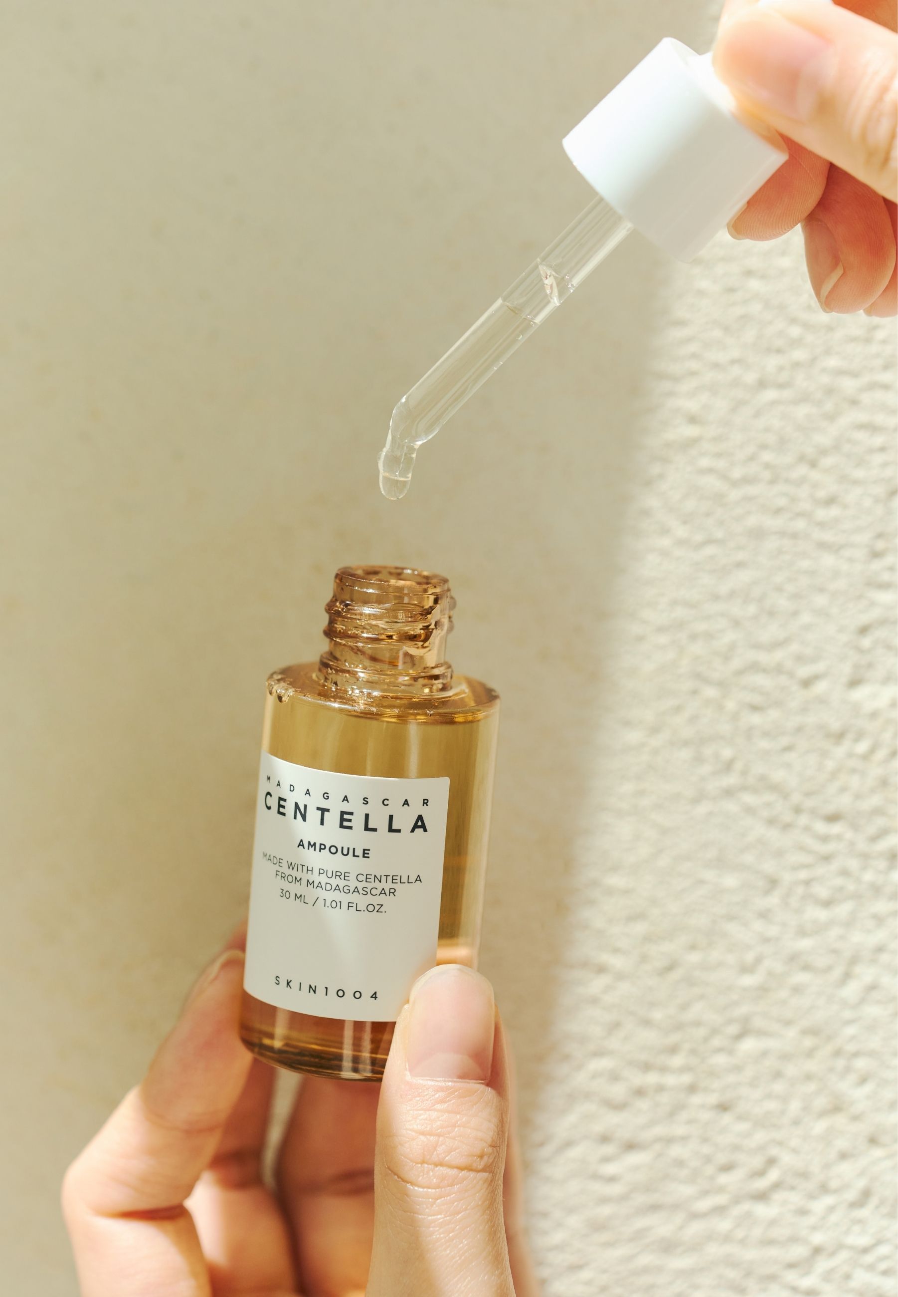 Handen houden een kleine fles met geelachtige vloeistof en pipet. Opschrift: MADAGASCAR CENTELLA AMPULE. SKIN1004.