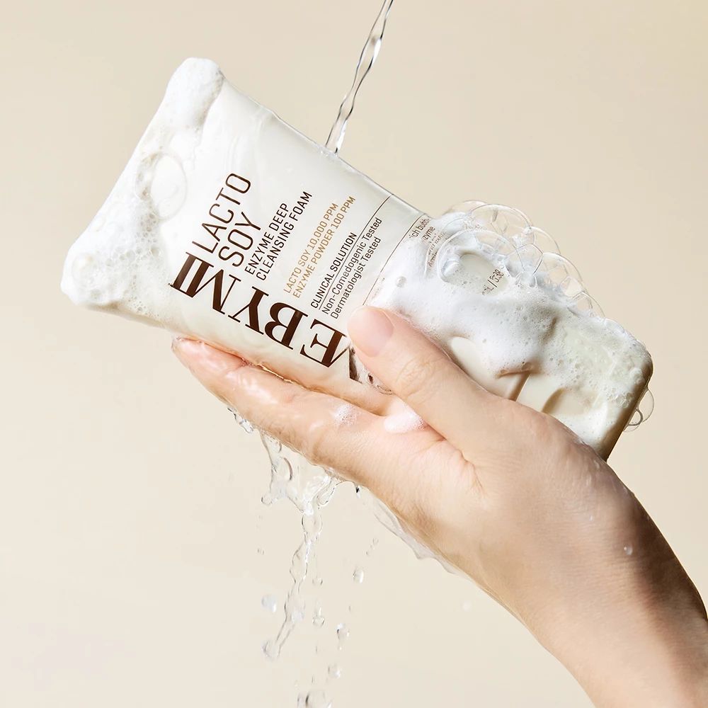 Une main tient un tube de mousse nettoyante blanche. De l'eau coule sur le tube et la mousse.