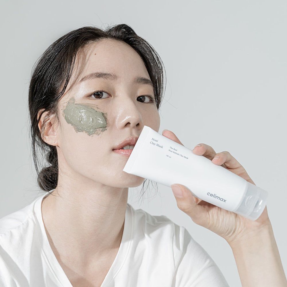 Une femme applique un masque à l'argile verte, tenant un tube CELIMAX "The Real Noni Refresh Clay Mask".