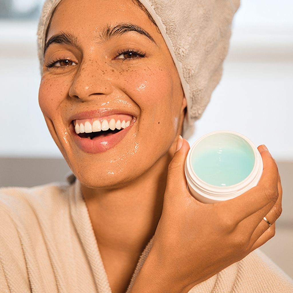 Vrouw met handdoek en badjas houdt open pot vast. Gel-gezichtsmasker zichtbaar. Gezicht glanst.