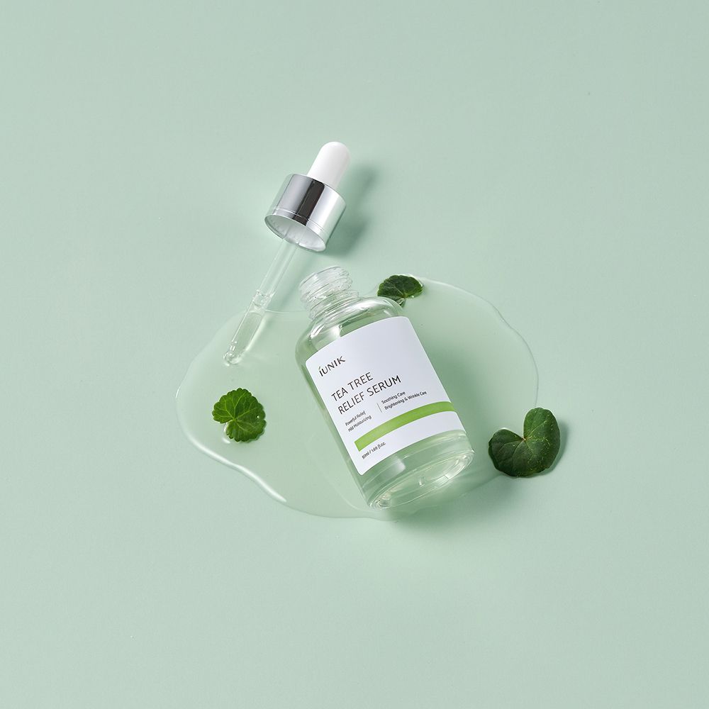 Flacon iUNIK Tea Tree Relief Serum avec compte-gouttes. Liquide sur surface avec feuilles. Fond vert clair.