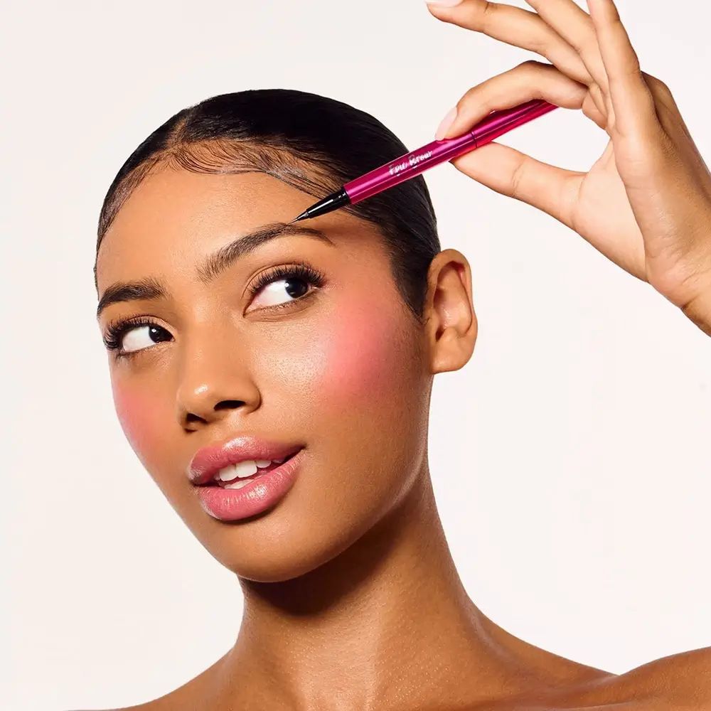 Hand houdt roze pen bij wenkbrauw. Zwarte punt. Gezicht met blush.