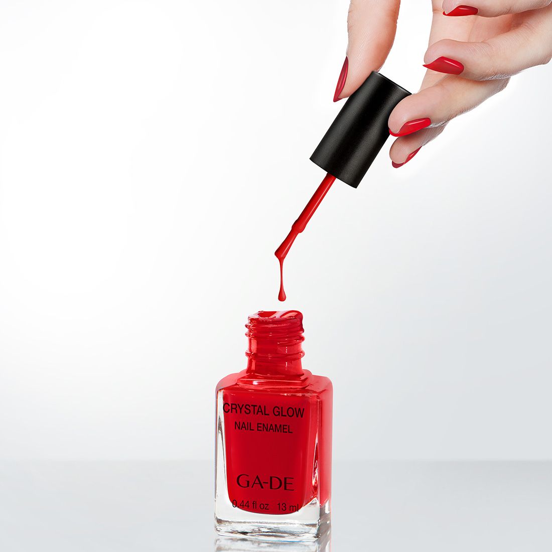 Rode nagellak in glazen fles, kwast met druppel. Opschrift: Crystal Glow Nail Enamel, GA-DE. Hand met gelakte nagels.