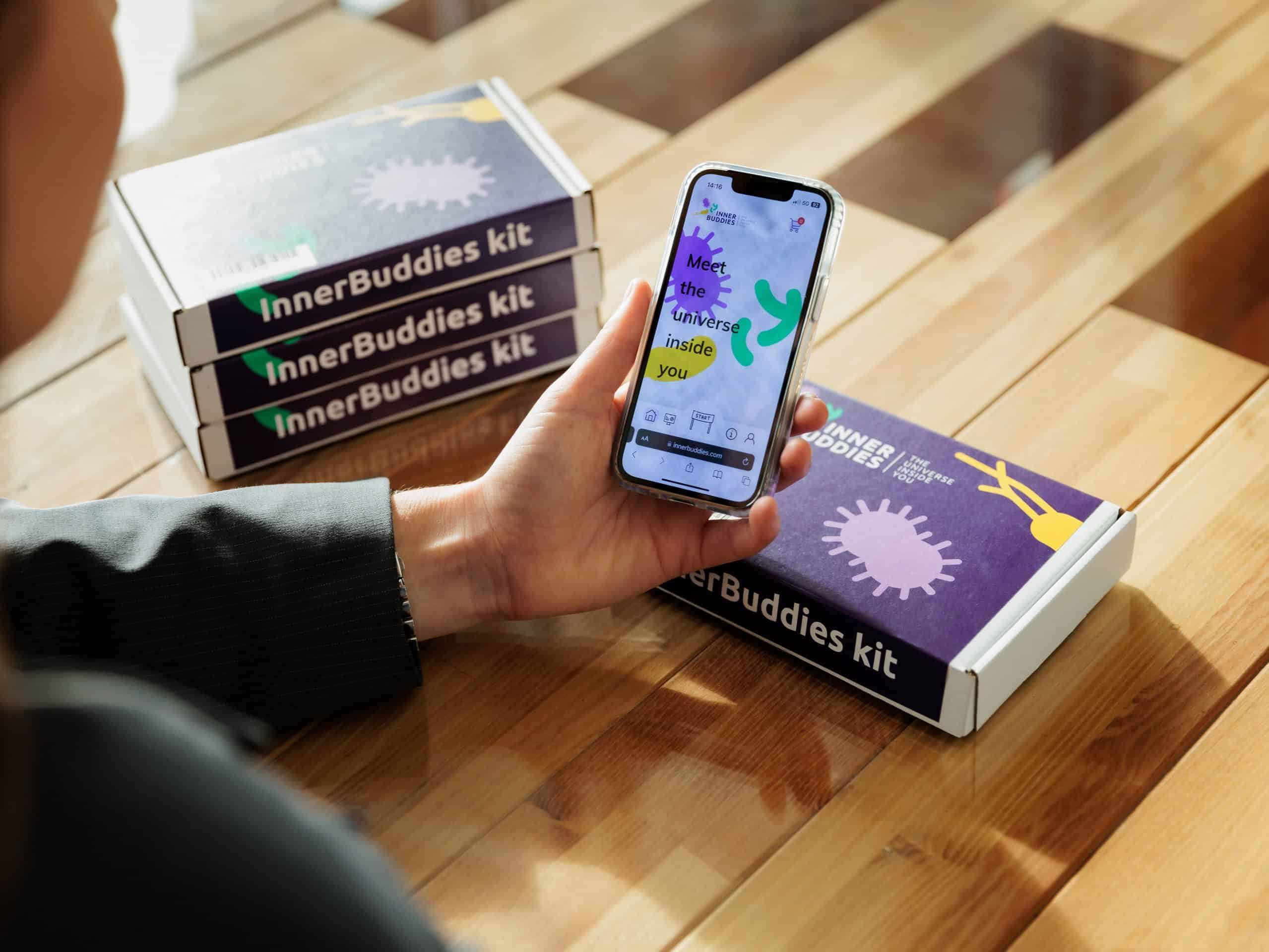 Drie gestapelde Inner Buddies testkit verpakkingen naast een persoon die een smartphone vasthoudt.