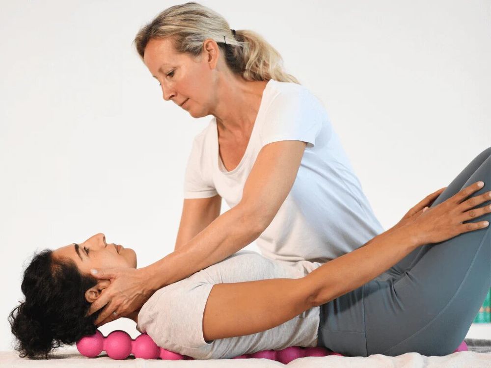 Vrouw ligt op rug, hoofd op roze SPINEFITTER®. Andere vrouw masseert haar nek.