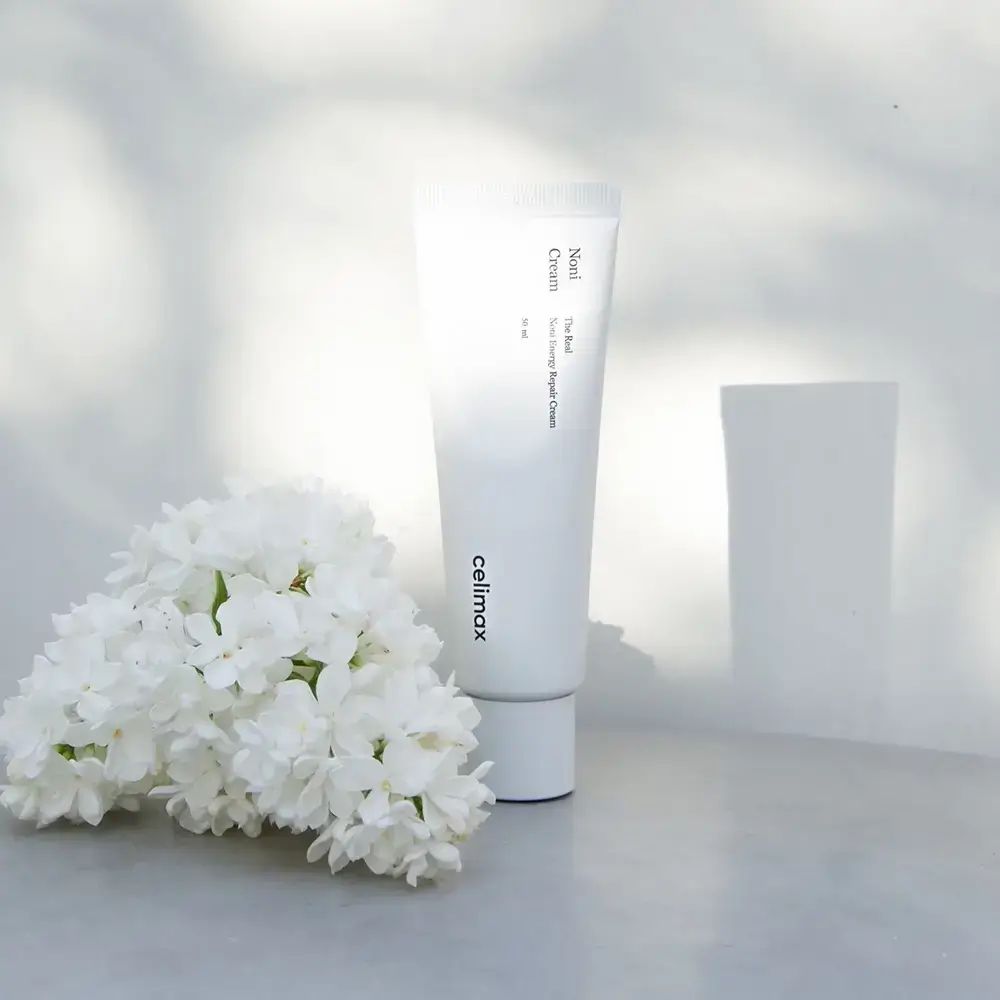 Witte tube met productnaam en logo. Witte bloemen en lichte achtergrond. Productnaam: Noni Cream.