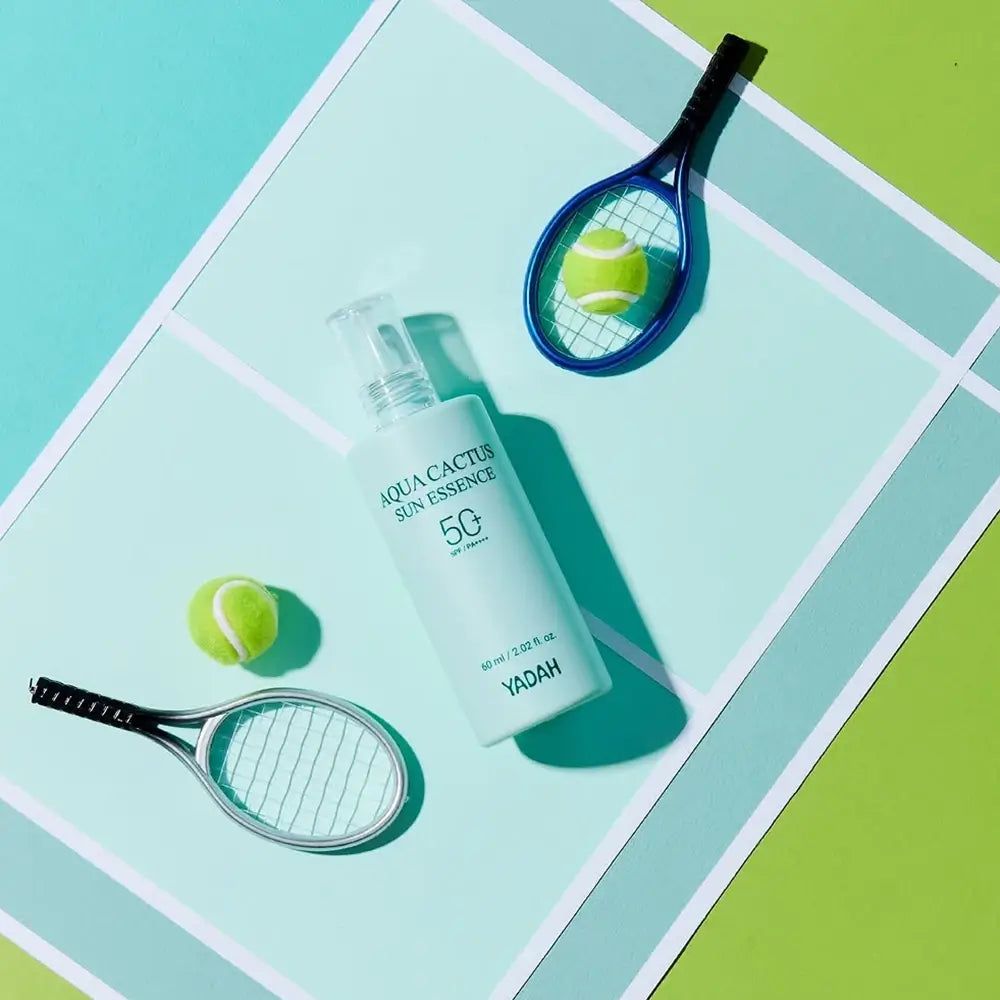 Lichtgroene fles met witte applicator. Opschrift: Aqua Cactus Sun Essence 50. Twee tennisballen en rackets ernaast.