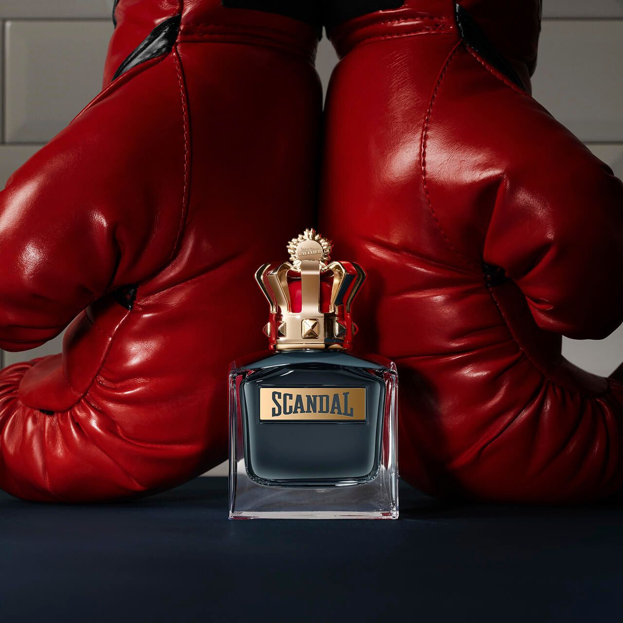 Flacon de parfum avec couronne dorée devant des gants de boxe rouges. Inscription "SCANDAL".