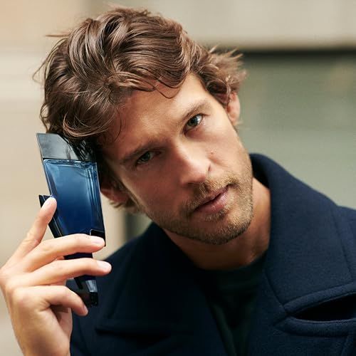 Een man houdt een blauwe fles in bliksemvorm vast. Hij draagt een donkere jas en kijkt in de camera.