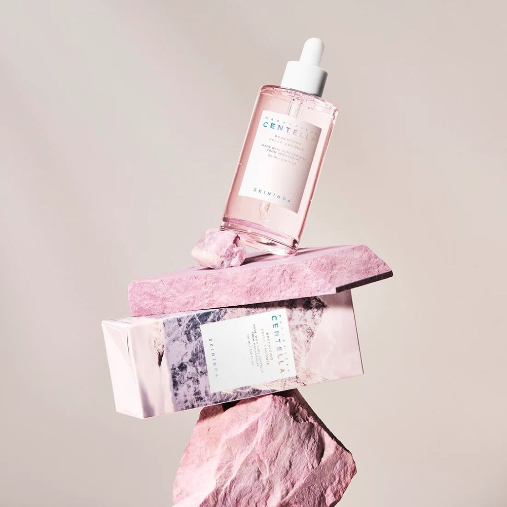 Serumfles op roze stenen. Roze verpakking eronder. Merk SKIN1004 zichtbaar.