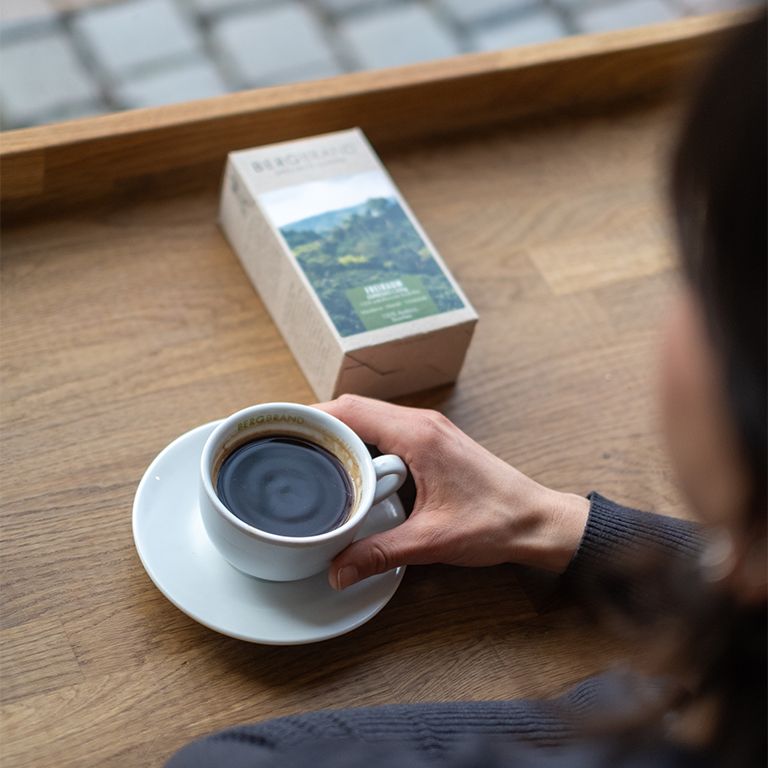 Personne tenant une tasse de café sur une soucoupe. À côté, un paquet de café. Sur une table en bois.