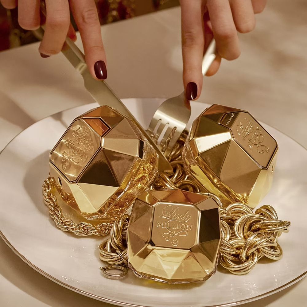 Trois flacons Lady Million dorés sur une assiette avec une chaîne dorée. Fourchette et couteau à côté.