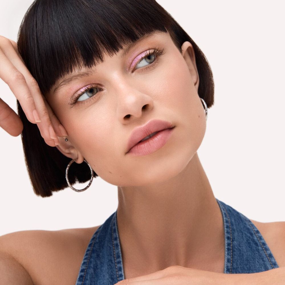 Femme avec fard à paupières rose. Elle porte une salopette en jean et des créoles argentées.