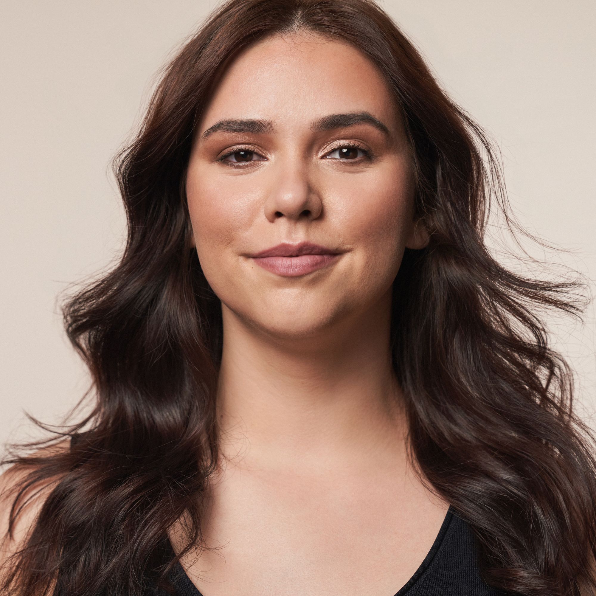 Portrait d'une femme aux longs cheveux bruns. Elle porte du maquillage, y compris du contouring. Elle sourit légèrement.