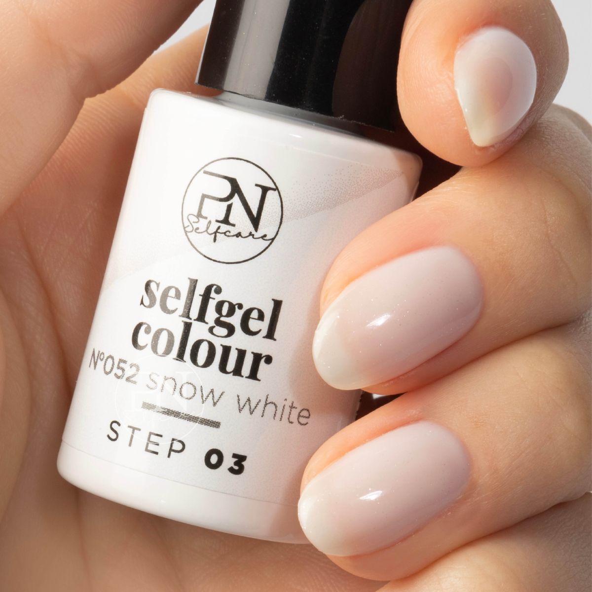 Une main tient un flacon de vernis à ongles. Inscriptions : PN Selfcare, selfgel colour, Snow White, STEP 03. Ongles vernis de couleur claire.