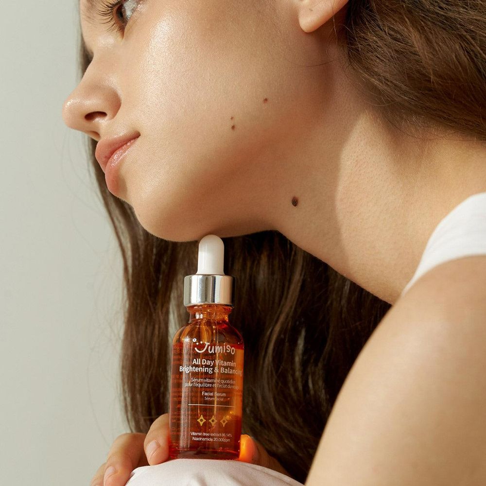 Vrouw houdt een flesje serum vast. Opschrift: JUMISO All Day Vitamin Brightening & Balancing. Gezicht in profiel.