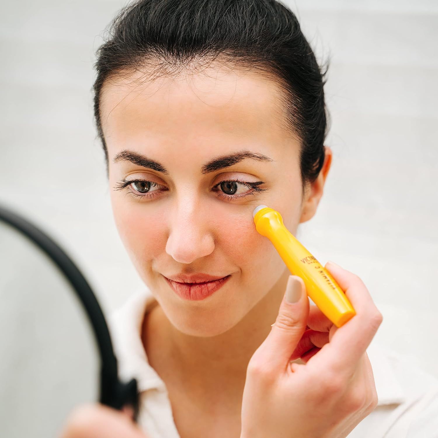 Vrouw brengt gele oogroller aan. Product wordt onder het oog aangebracht. Spiegel op de achtergrond.