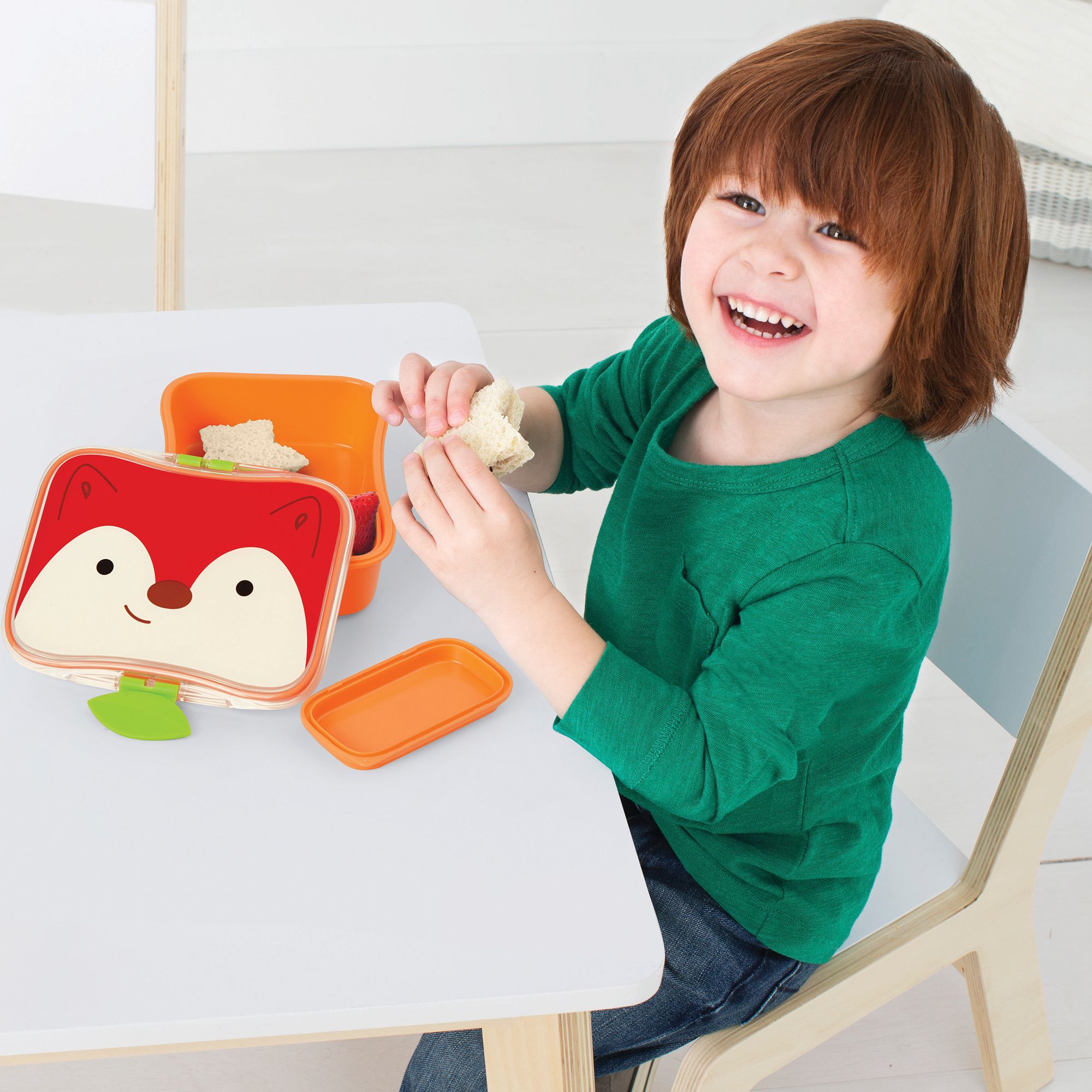 Kind opent lunchbox met vossendesign aan tafel. Klein bakje en sandwich zichtbaar.