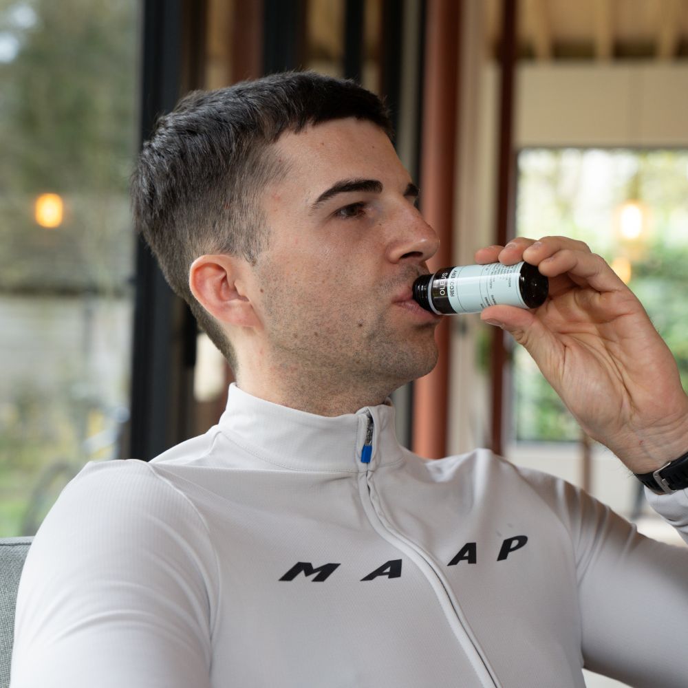 Persoon drinkt uit een flesje met zwarte dop en etiket. Op het etiket staat: OLEUS, Recovery.