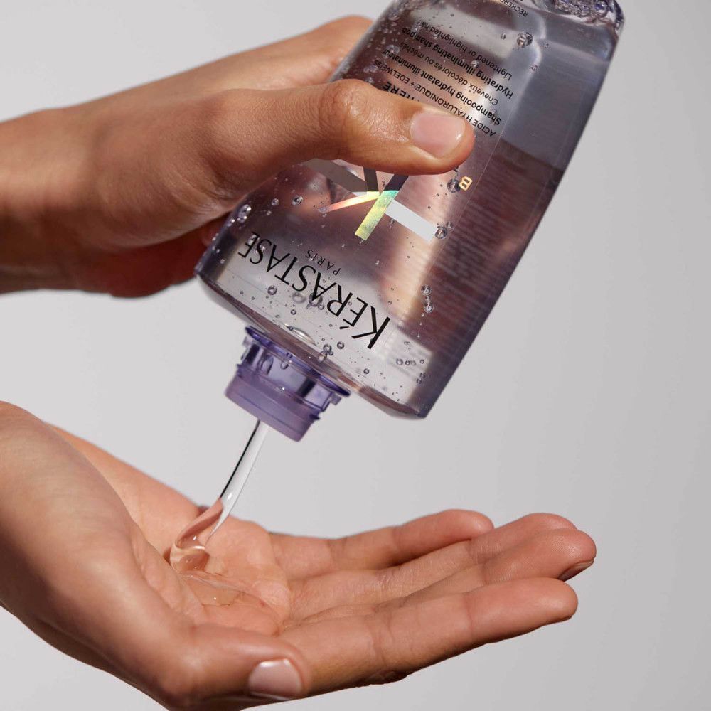 Kérastase Blond Absolu Shampoo: handen gieten shampoo in een handpalm. Paarse transparante fles zichtbaar.