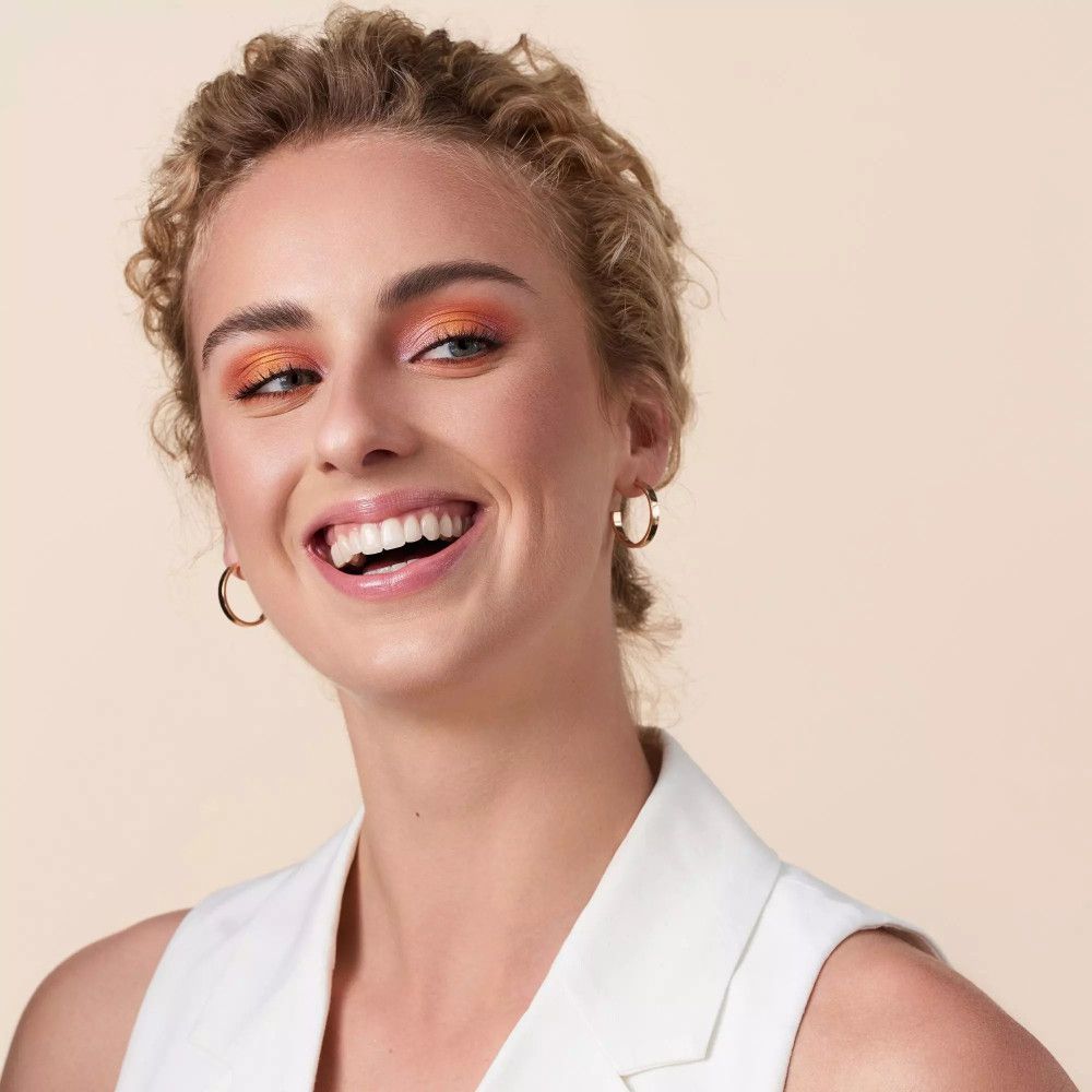 Portret van een lachende vrouw met blonde krullen en gouden oorringen.