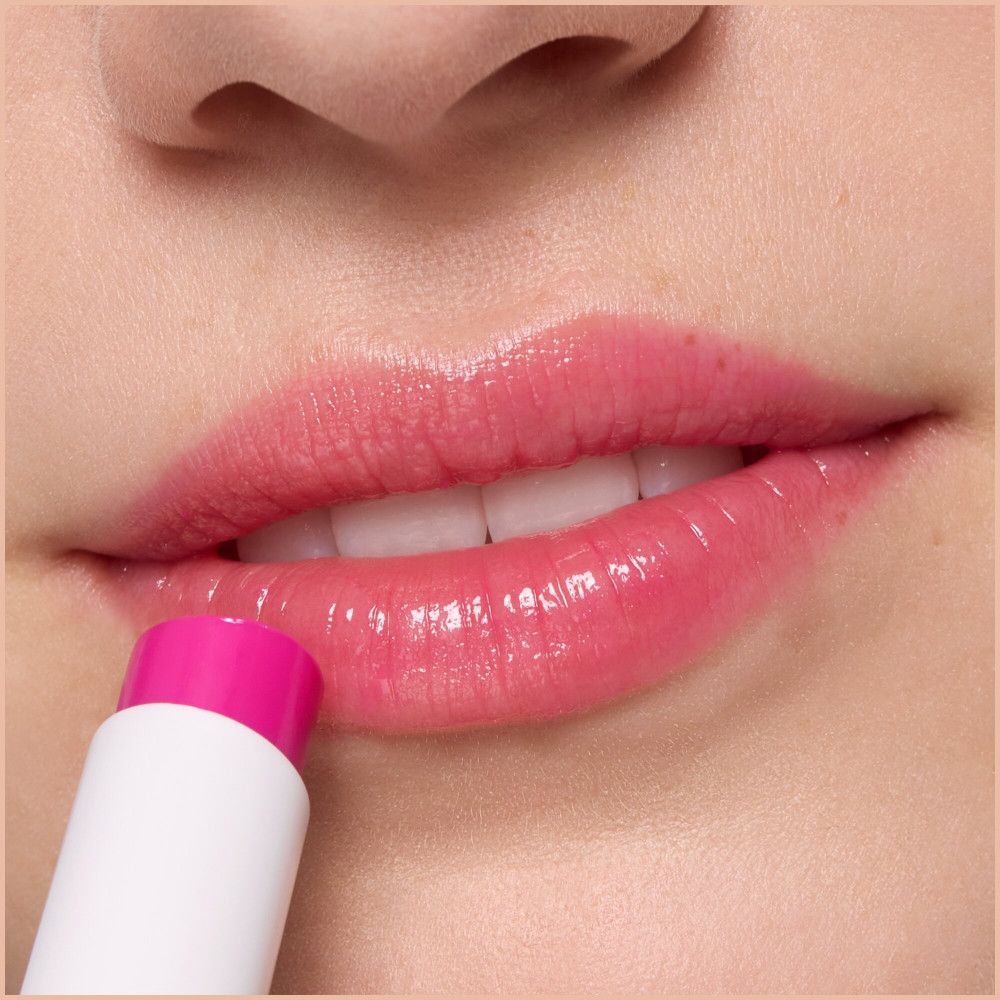 Gros plan sur des lèvres recouvertes de baume à lèvres rose. Le baume est dans un tube blanc.