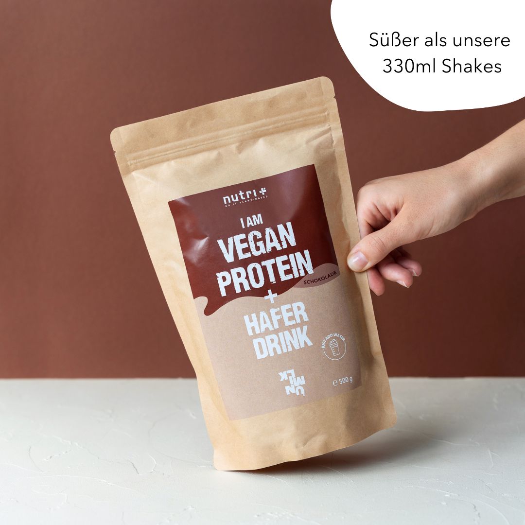 Beige zak met 'I AM VEGAN PROTEIN + HAFER DRINK'. Bruine opdruk. Hand houdt de zak vast. Tekst: 'Süßer als unsere 330ml Shakes'.