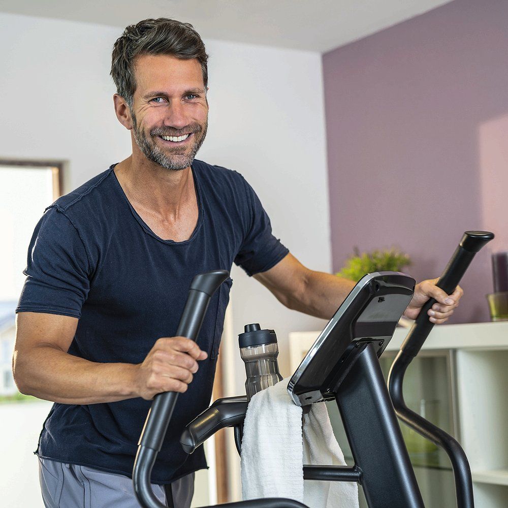 Homme s'entraînant sur un elliptique. Bouteille d'eau et serviette. Dans un salon. Sourit à la caméra.