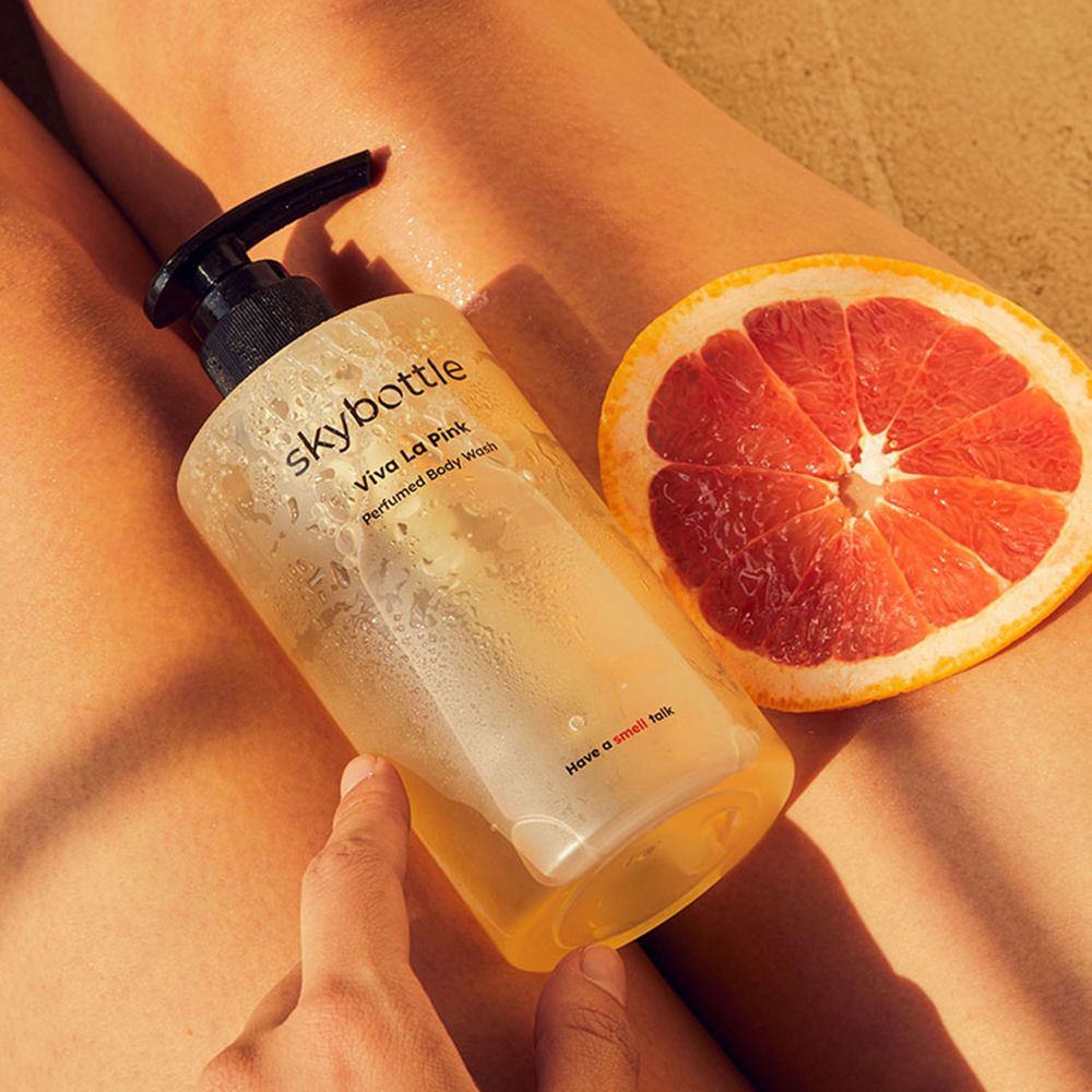 Een persoon houdt een fles douchegel vast naast een halve grapefruit. Benen op de achtergrond.