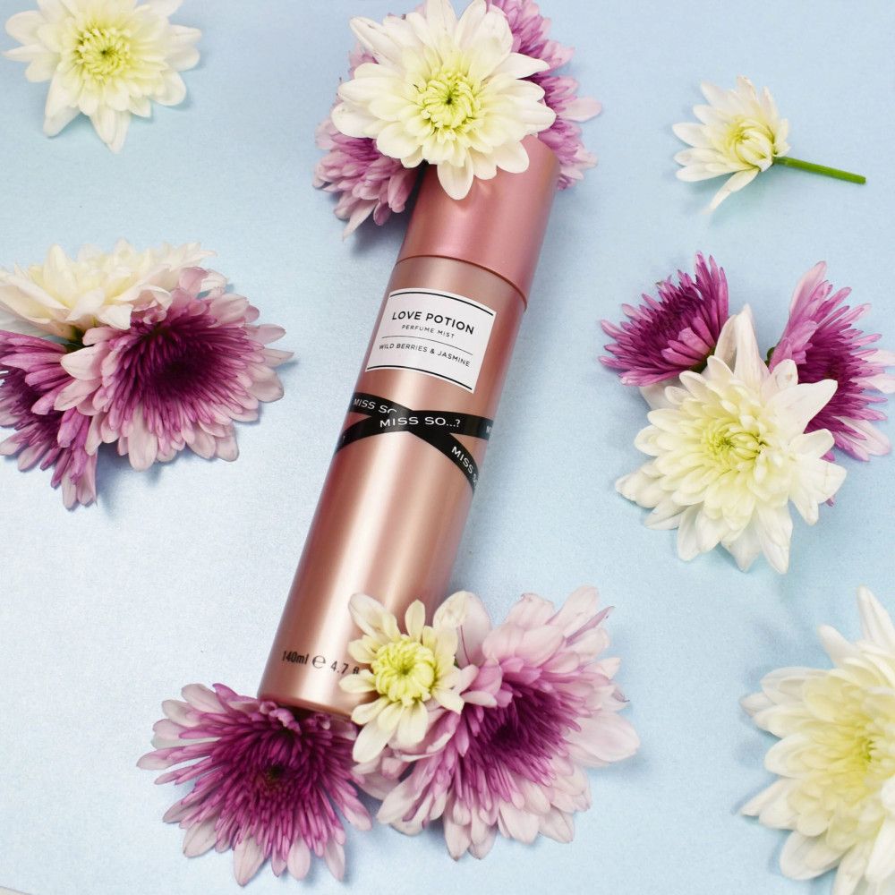 Vaporisateur corporel rose avec étiquette blanche. Inscription : Love Potion, Wild Berries & Jasmine, Miss So...?. Entouré de fleurs.